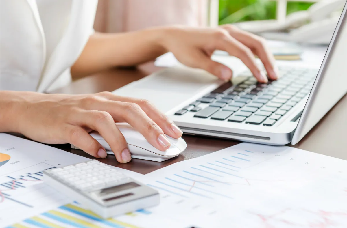 Hands using a laptop and mouse beside printed financial charts