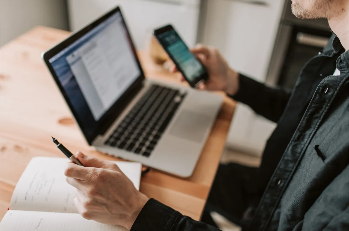 Person using a phone and a laptop while taking notes