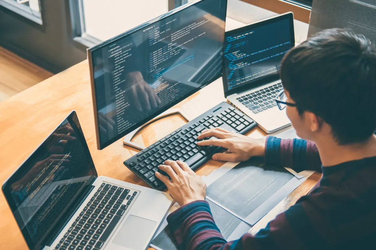 Software developer writing code on a laptop and two monitors